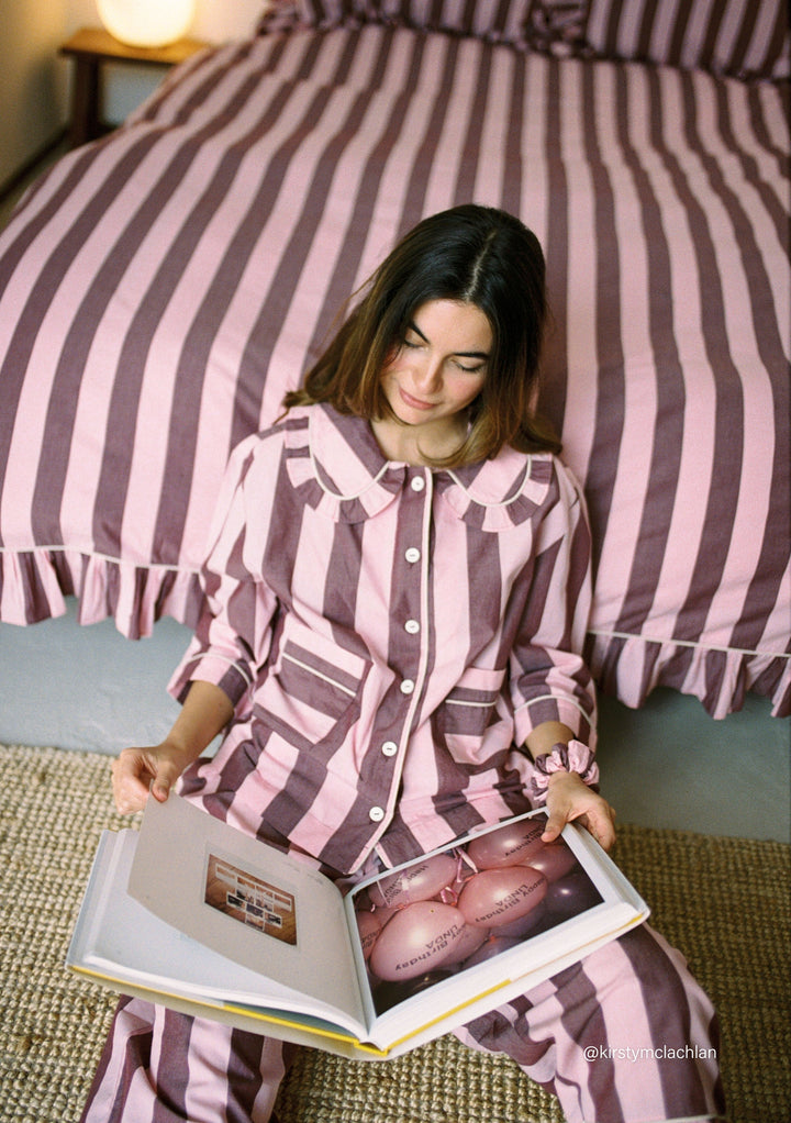 Woman sitting on carpet reading book, wearing TBCo Cropped Cotton Pyjamas in Pink & Burgundy Stripe matching bedding pattern