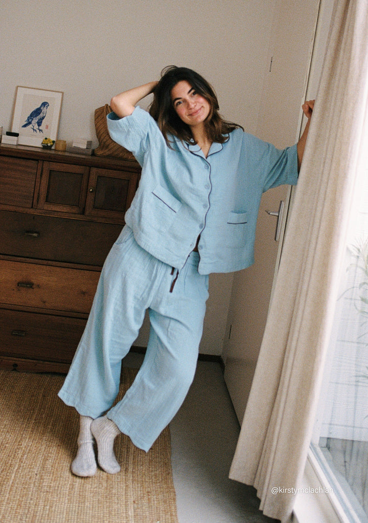 Woman stretching by curtain wearing TBCo Cropped Cotton Pyjamas in Blue with brown contrast piping in cozy room