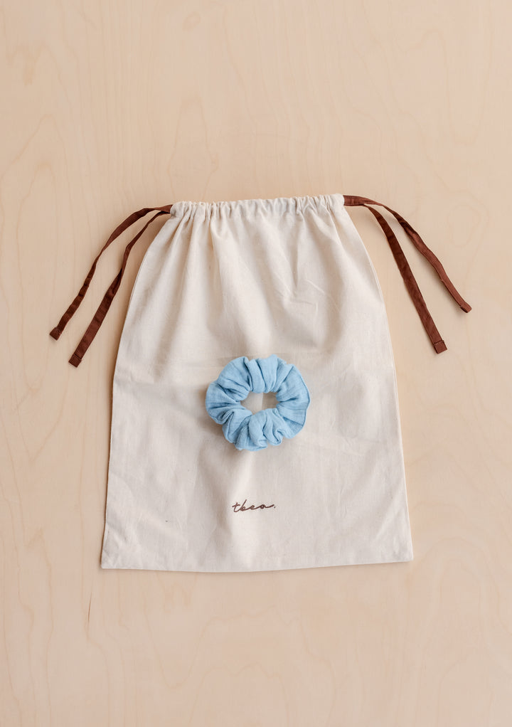 Blue scrunchie resting on cream drawstring bag with brown ties, styled with TBCo Cropped Cotton Pyjamas in Blue packaging