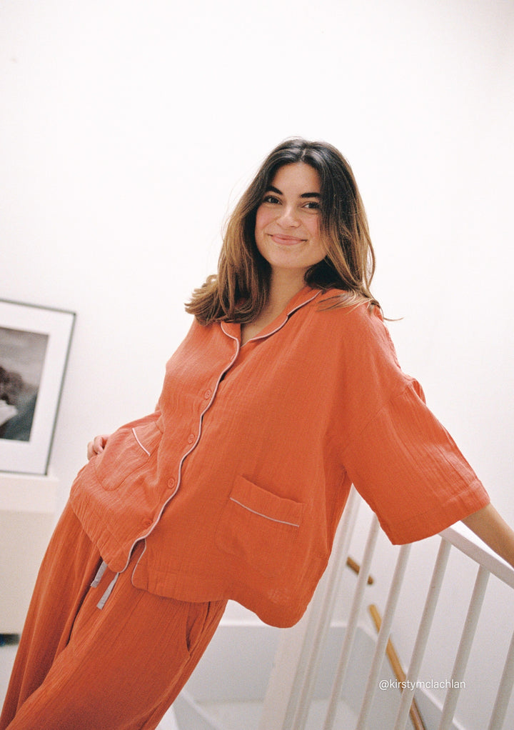 Woman wearing TBCo Cotton Pyjamas in Rust with lilac piping, relaxed fit, and button-up top smiling indoors on stairs
