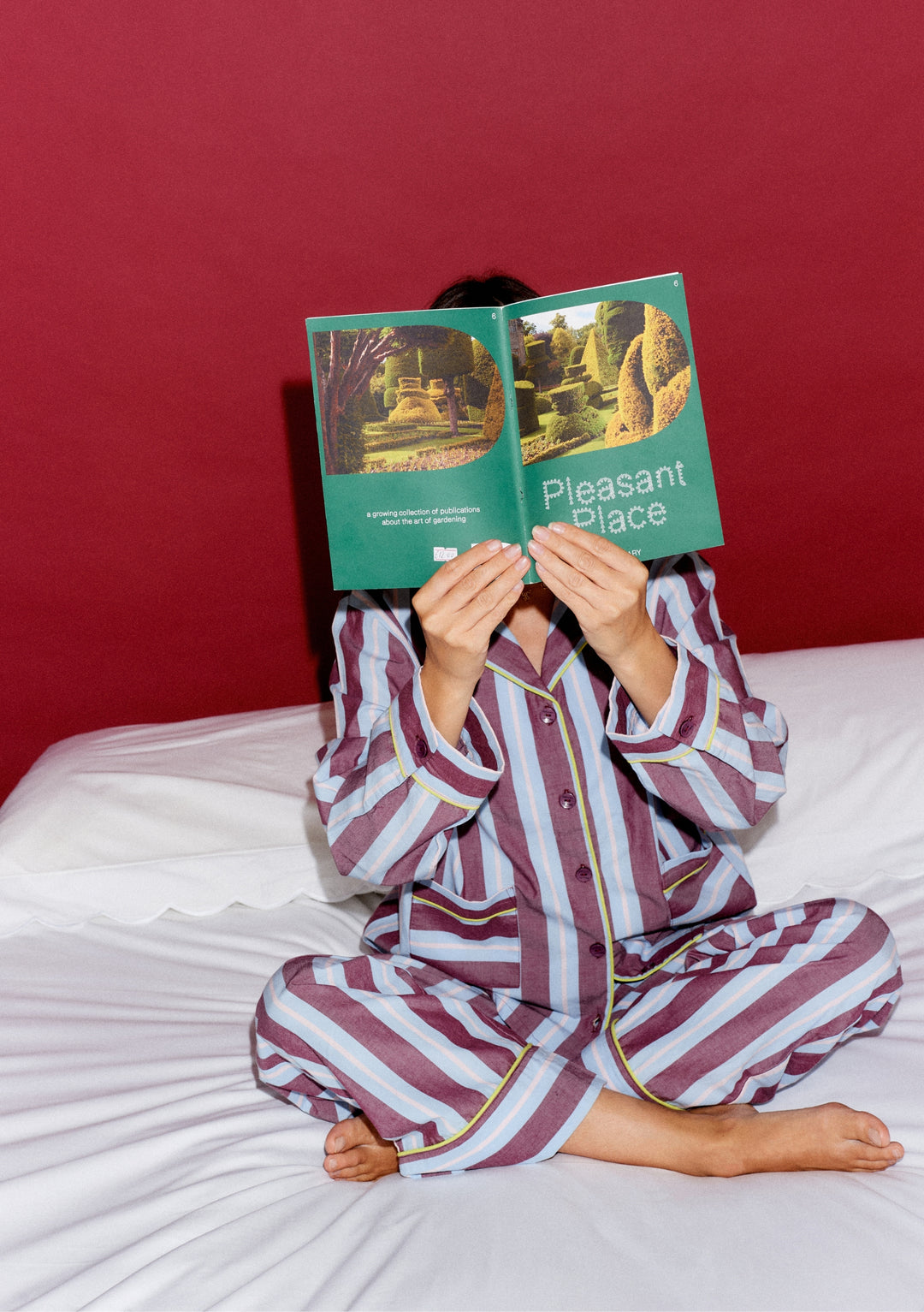 Person in TBCo x Glassette Cotton Pyjamas in Berry Stripe, seated cross-legged on a bed, holding a green booklet titled Pleasant Place in front of their face.