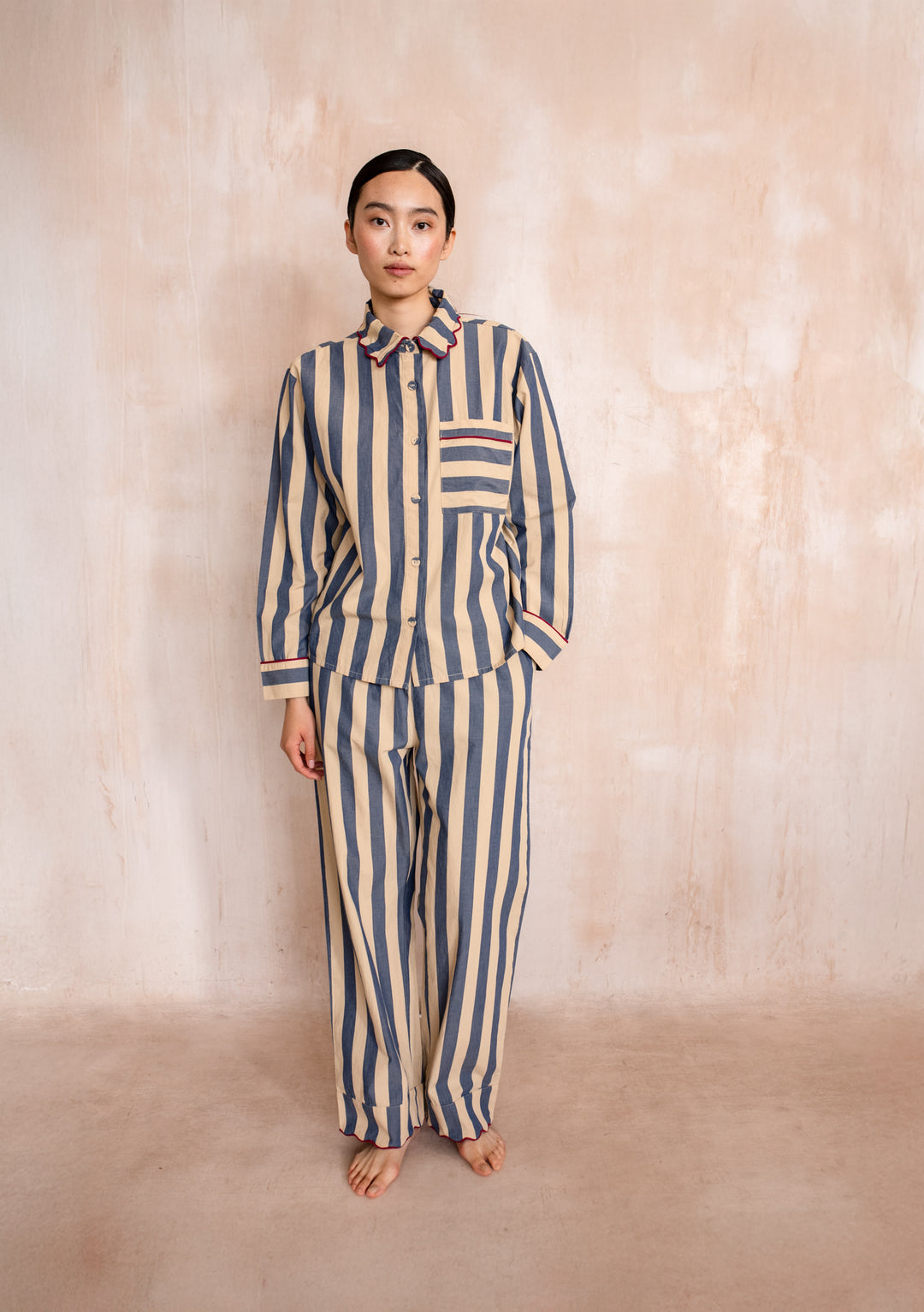 Model in a TBCo Cotton Pyjamas in Navy Stripe with scalloped collar, relaxed fit, showcasing modern sleepwear style against a textured wall.