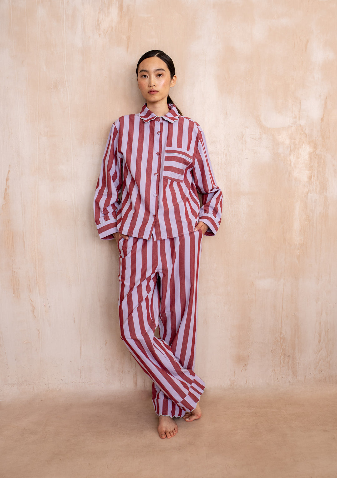Model in a TBCo Cotton Pyjamas in Lilac Stripe with scalloped collar, relaxed fit, showcasing modern sleepwear style against a textured wall.