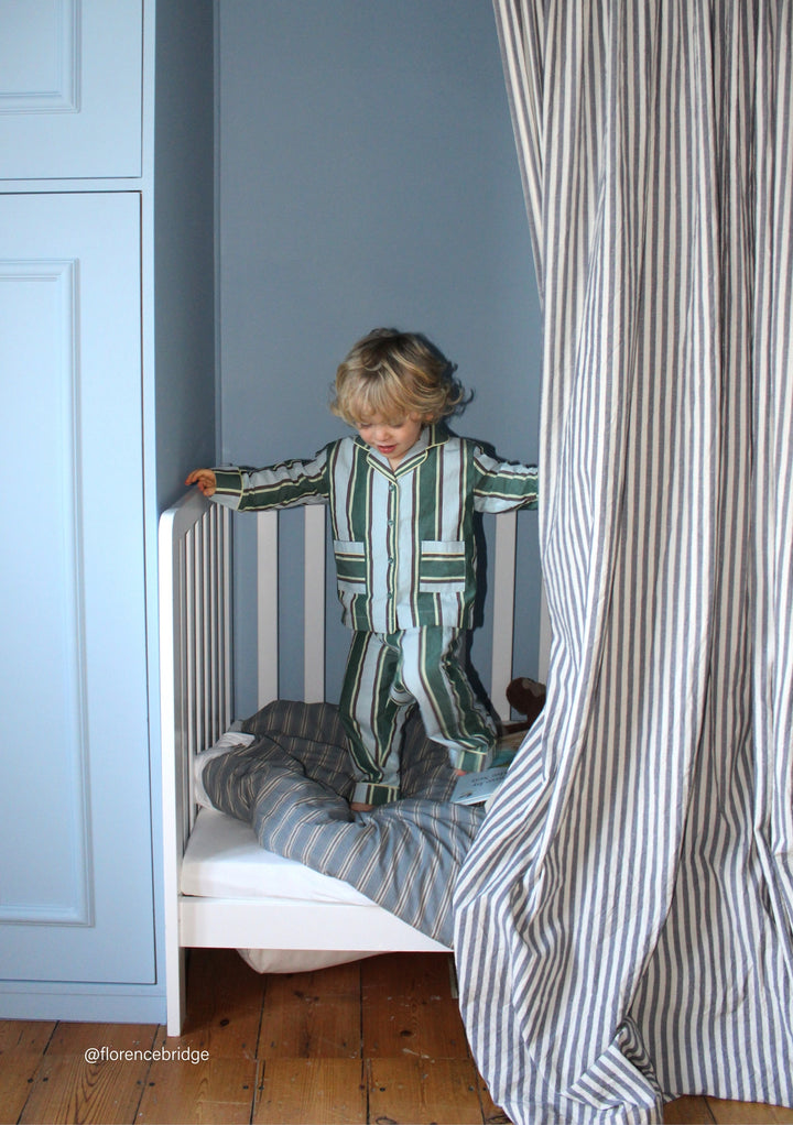 Child wearing TBCo Cotton Kids Pyjamas in Blue Stripe standing on white bed with striped bedding and curtain nearby