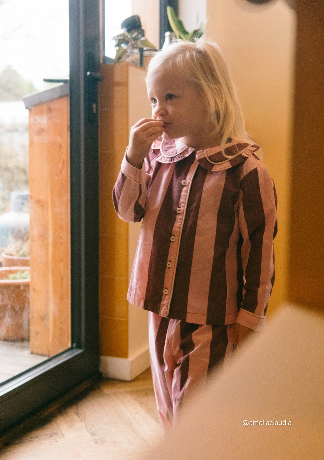 Child wearing TBCo Cotton Kids Pyjamas in Pink & Burgundy Stripe standing by glass door in a cosy indoor setting