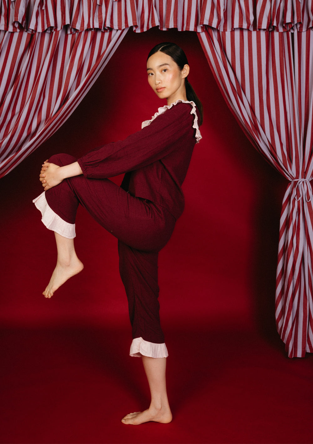 Model in TBCo Cotton Pyjamas in Burgundy Ruffle with pink trim, seated against a red backdrop, alongside  striped fabric curtain accent.