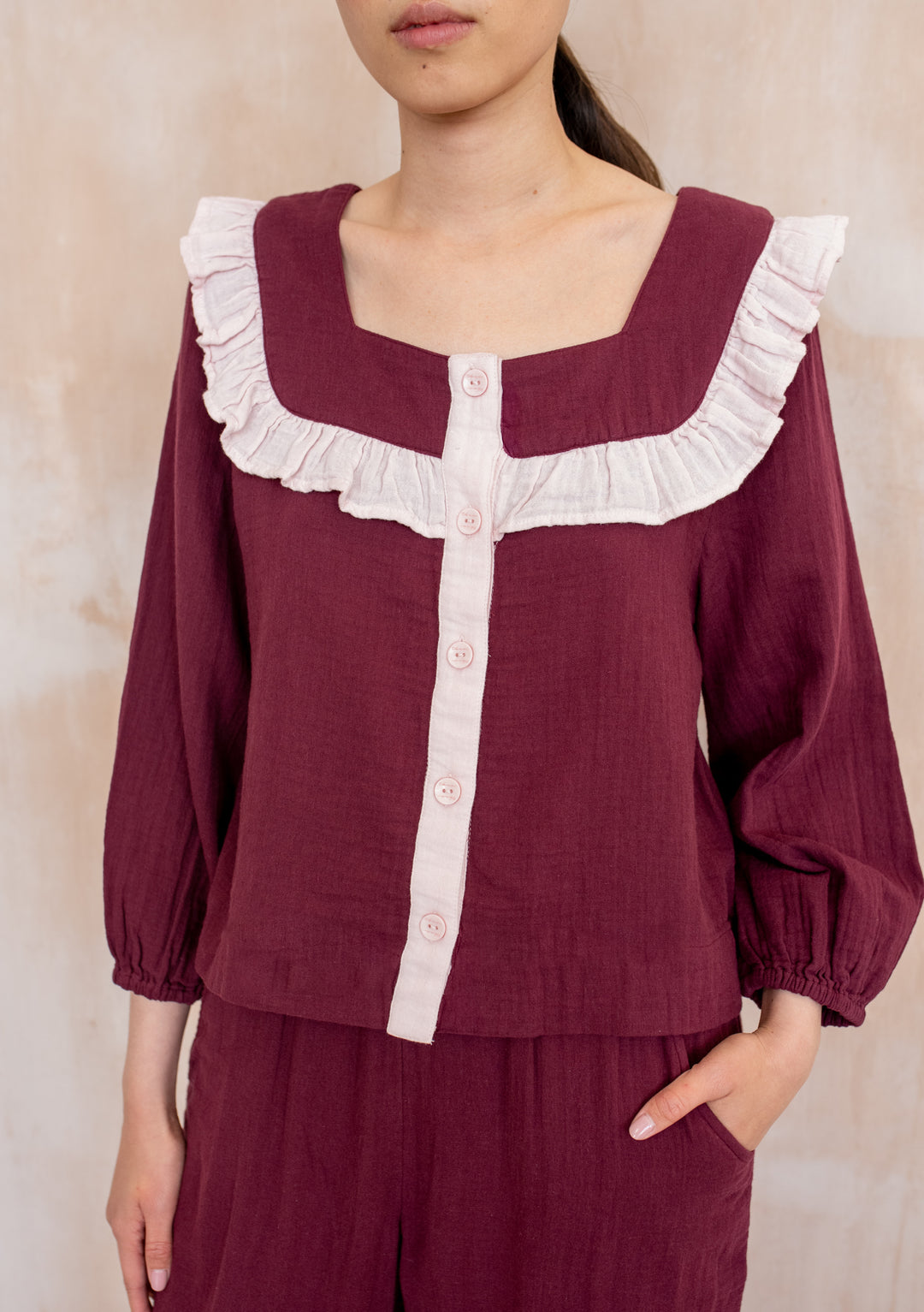 Person wearing TBCo Cotton Pyjamas Burgundy Ruffle with square neckline, pink ruffle trim, and buttons, featuring billowed sleeves, elastic cuffs, and waist-high bottoms with pockets.