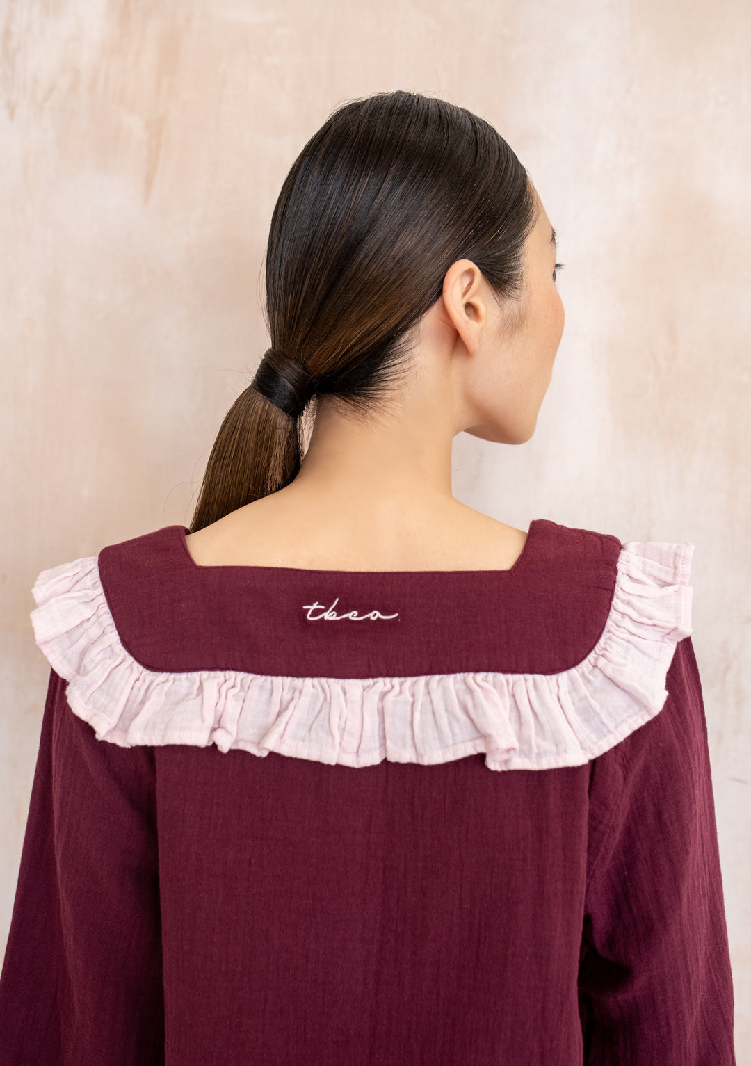 Back view of TBCo Cotton Pyjamas Burgundy Ruffle, featuring a sailor-style collar with pink ruffle and cursive TBCo embroidery, worn by a woman with a low ponytail.