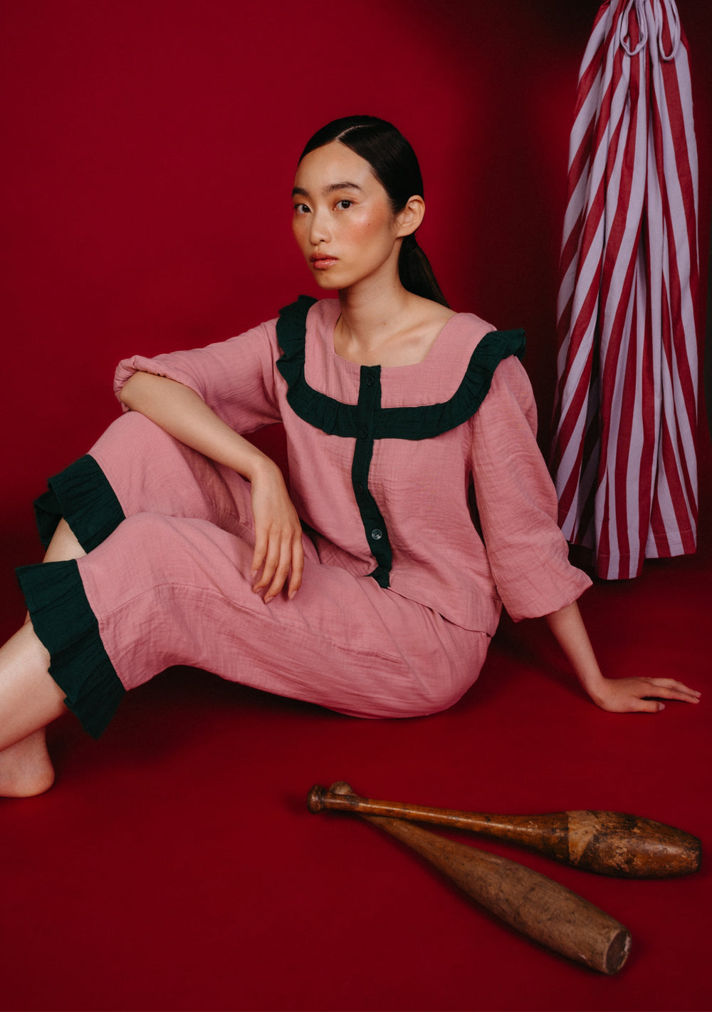 Woman in TBCo Cotton Pyjamas in Pink Ruffle with green trim, seated against a red backdrop, alongside wooden clubs and striped fabric accent.