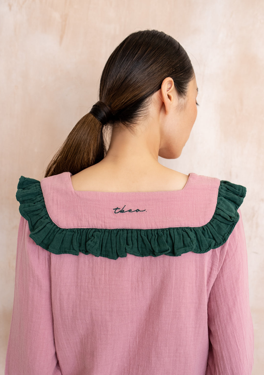 Back view of TBCo Cotton Pyjamas in Pink Ruffle, featuring a sailor-style collar with green ruffle and cursive TBCo embroidery, worn by a woman with a low ponytail.