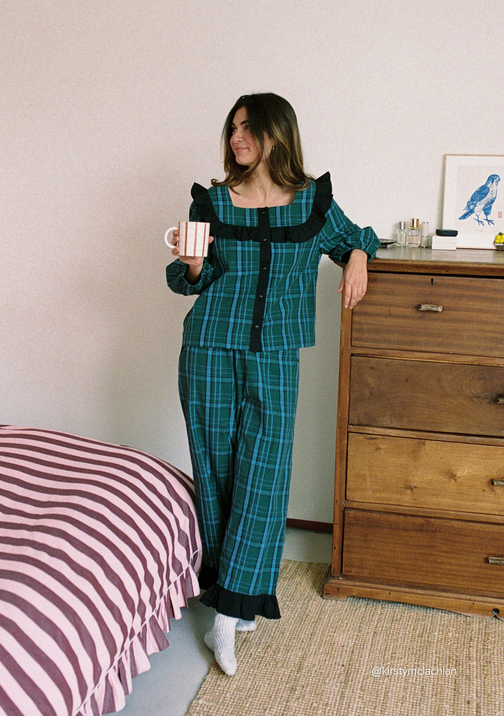 Woman wearing TBCo Cropped Cotton Pyjamas in Green Tartan with ruffle collar holding a mug by a wooden dresser
