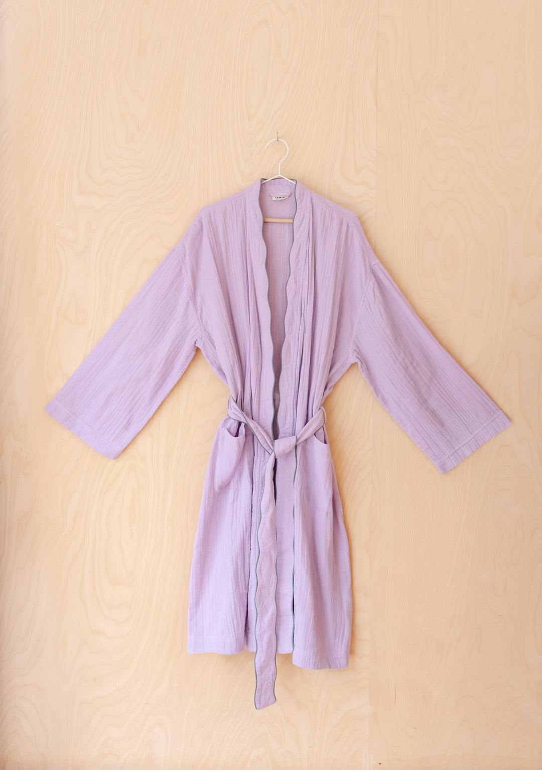 TBCo Cotton Robe in Lilac hanging against wooden wall, portrait orientation gallery position 4 with scalloped edges detail