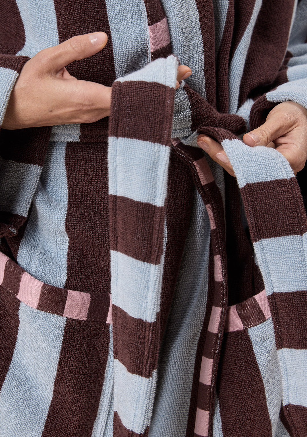 Close-up of hands adjusting the belt on TBCo Terry Cotton Bath Robe in Blue Stripe featuring soft textured fabric and colour block design