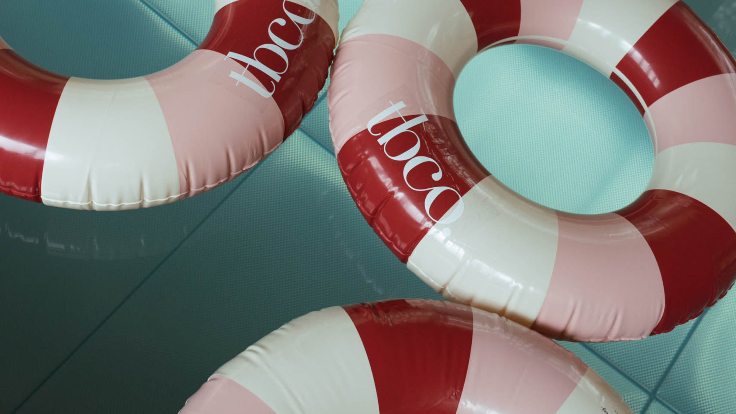 Three inflatable TBCo branded  rings with red, white, and pink stripes in a blue pool