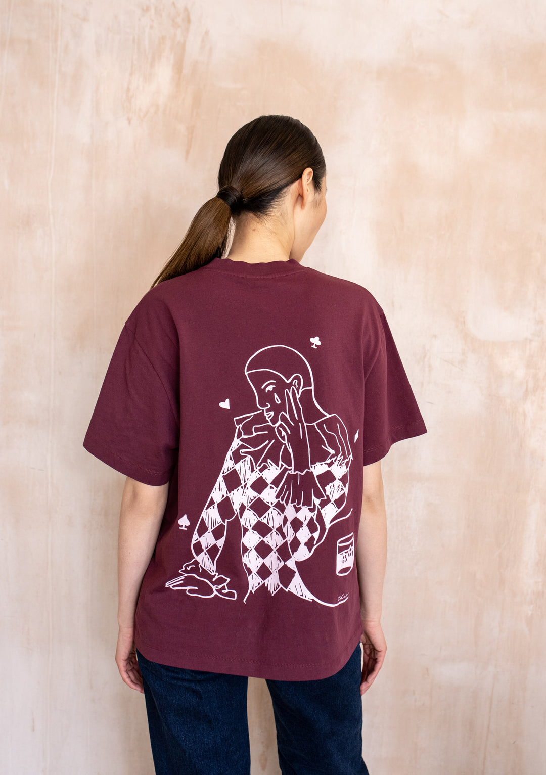 Back view of a model wearing the TBCo Cotton T-Shirt in Harlequin, showcasing a harlequin graphic with playing-card symbols on its burgundy back.