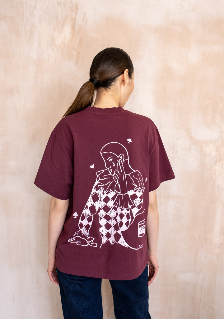 Back view of a model wearing the TBCo Cotton T-Shirt in Harlequin, showcasing a harlequin graphic with playing-card symbols on its burgundy back.