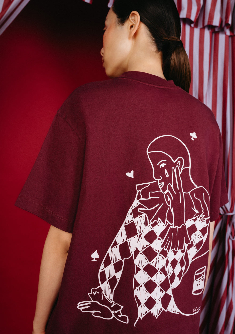 Back view of a model wearing the TBCo Cotton T-Shirt in Harlequin, showcasing a harlequin graphic with playing-card symbols on its burgundy back.