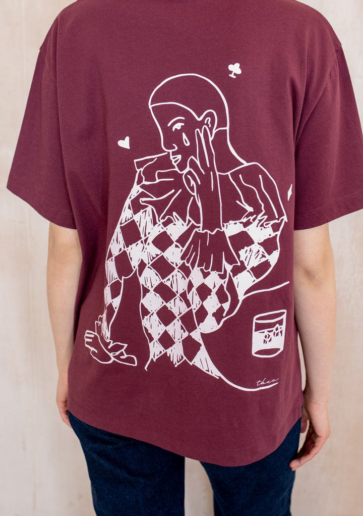 Back view of a model wearing the TBCo Cotton T-Shirt in Harlequin, showcasing a harlequin graphic with playing-card symbols on its burgundy back.