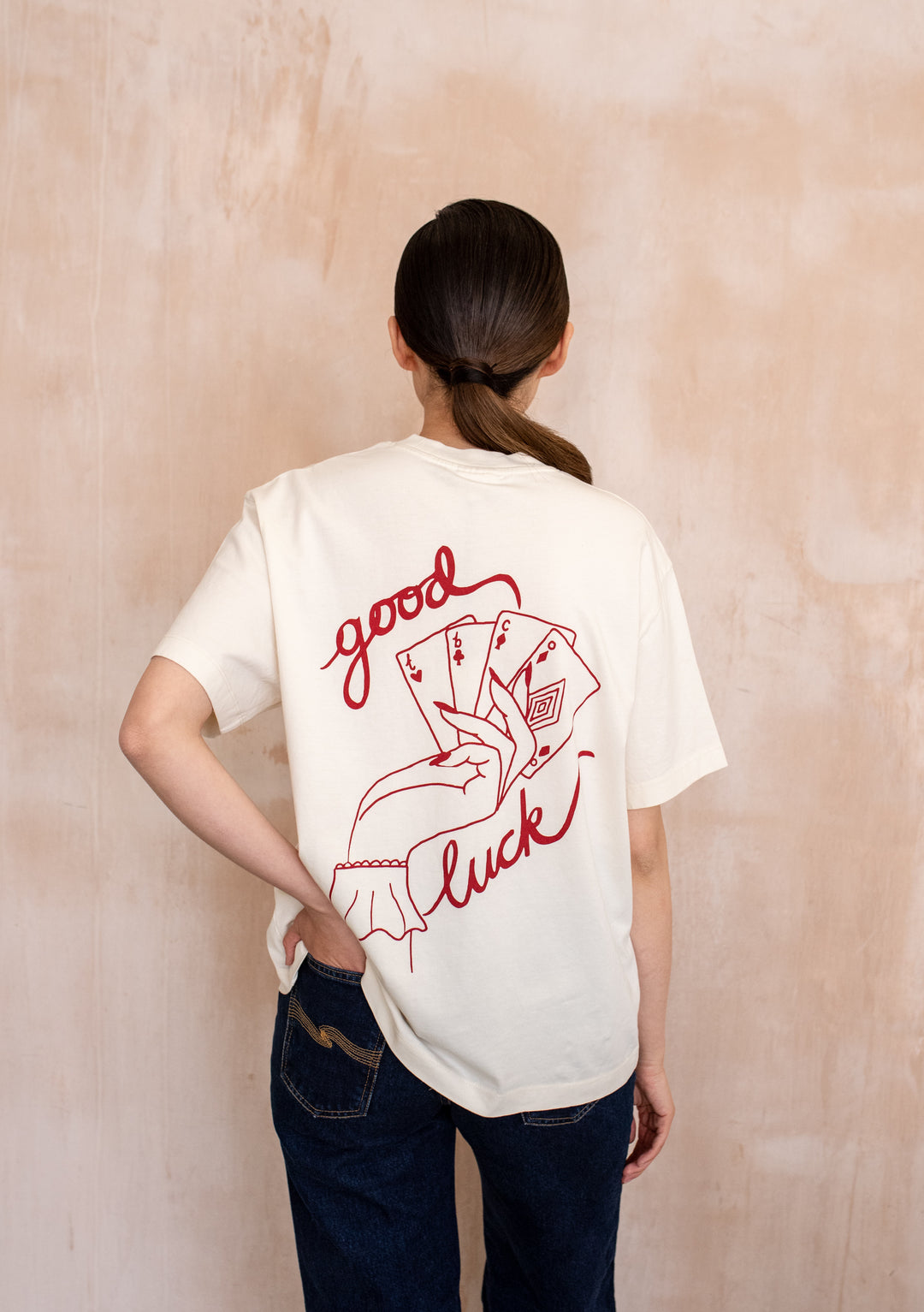 Model wearing the TBCo Cotton T-Shirt in Good Luck Cards, featuring a playing card graphic and good luck text, paired with dark blue jeans.