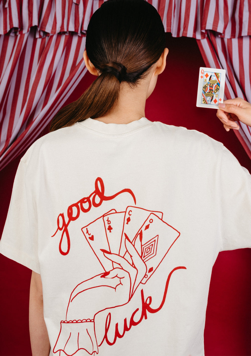 Model wearing the TBCo Cotton T-Shirt in Good Luck Cards while holding up a Queen of Hearts playing card, featuring a playing card graphic and good luck text, paired with dark blue jeans.