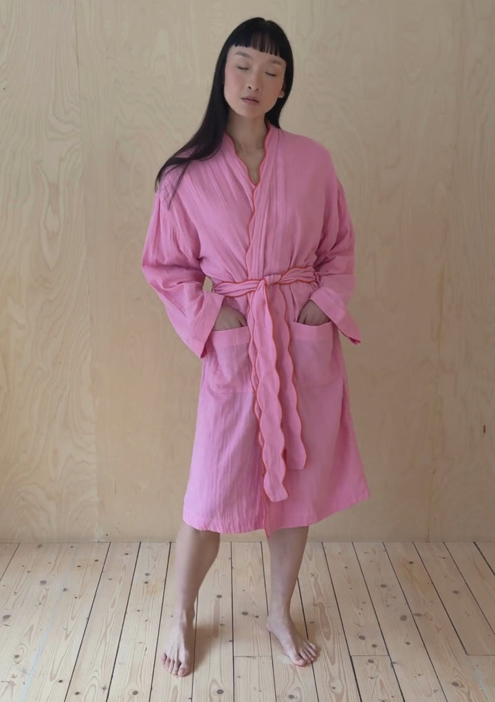 A model poses against a neutral ply backdrop, wearing a TBCo pink cotton robe.